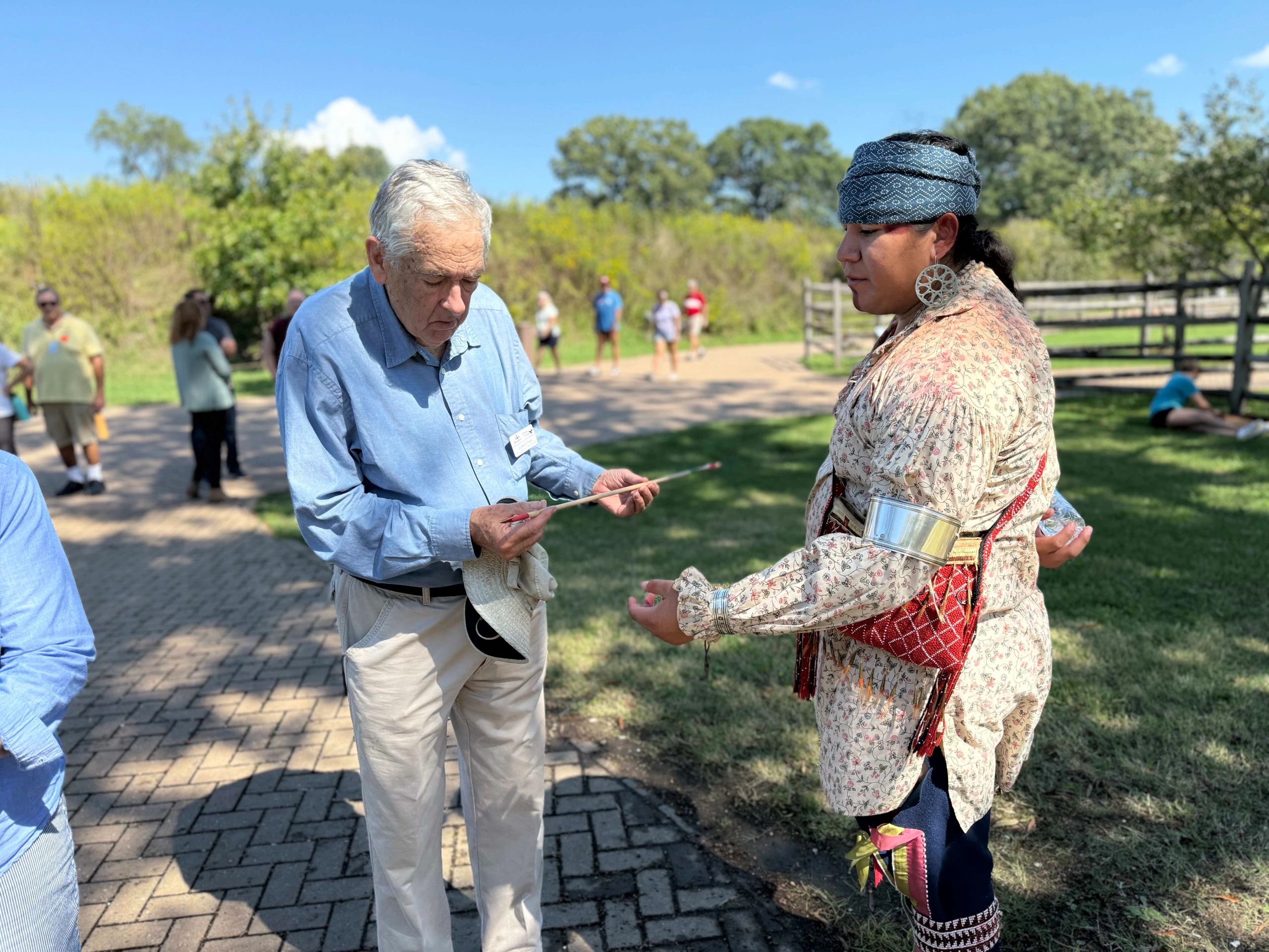 An elderly man interacts with a Native American man in traditional attire outdoors.